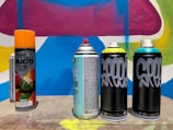 Several spray paint cans are arranged on a concrete surface in front of a colorful graffiti wall with abstract shapes in blue, pink, yellow, and white. The cans vary in design, with some featuring bold text and vibrant colors.