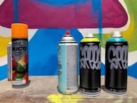 Several spray paint cans are arranged on a concrete surface in front of a colorful graffiti wall with abstract shapes in blue, pink, yellow, and white. The cans vary in design, with some featuring bold text and vibrant colors.