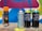 Several spray paint cans are arranged on a concrete surface in front of a colorful graffiti wall with abstract shapes in blue, pink, yellow, and white. The cans vary in design, with some featuring bold text and vibrant colors.