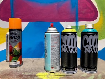 Several spray paint cans are arranged on a concrete surface in front of a colorful graffiti wall with abstract shapes in blue, pink, yellow, and white. The cans vary in design, with some featuring bold text and vibrant colors.