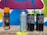 Several spray paint cans are arranged on a concrete surface in front of a colorful graffiti wall with abstract shapes in blue, pink, yellow, and white. The cans vary in design, with some featuring bold text and vibrant colors.