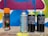 Several spray paint cans are arranged on a concrete surface in front of a colorful graffiti wall with abstract shapes in blue, pink, yellow, and white. The cans vary in design, with some featuring bold text and vibrant colors.