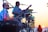 A musician playing drums on stage during a sunset, with a microphone in front of him. Another musician with a guitar is visible in the background, silhouetted by the setting sun. The scene conveys a lively and relaxed outdoor performance.