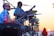 A musician playing drums on stage during a sunset, with a microphone in front of him. Another musician with a guitar is visible in the background, silhouetted by the setting sun. The scene conveys a lively and relaxed outdoor performance.