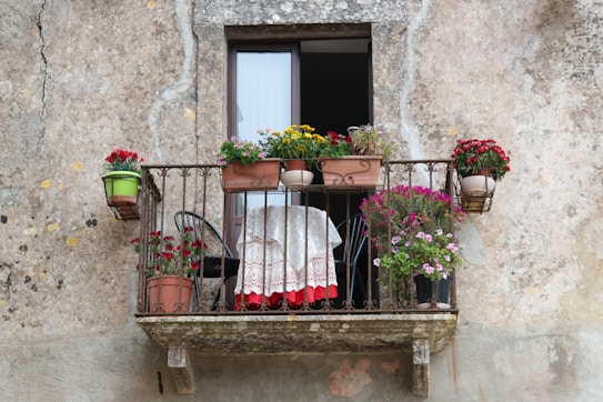 A small, rustic balcony with a variety of potted flowers and plants arranged on a metal railing. There's a small table with a delicate lace cloth, bordered in red, accompanied by a black chair. The background shows a textured wall, suggesting an aged, weathered building.
