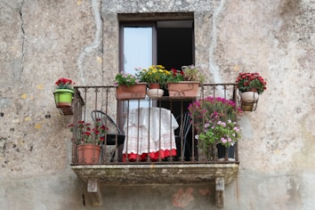 A small, rustic balcony with a variety of potted flowers and plants arranged on a metal railing. There's a small table with a delicate lace cloth, bordered in red, accompanied by a black chair. The background shows a textured wall, suggesting an aged, weathered building.