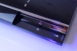 A sleek, black gaming console with a glossy finish is shown. The console features a disc drive, the PlayStation logo, and illuminated icons. The Sony branding is visible on the lower part, and the console is highlighted with a soft blue lighting accentuating its modern design.