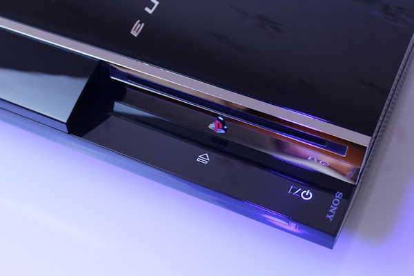 A sleek, black gaming console with a glossy finish is shown. The console features a disc drive, the PlayStation logo, and illuminated icons. The Sony branding is visible on the lower part, and the console is highlighted with a soft blue lighting accentuating its modern design.