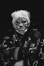 An elderly person with light hair has their face painted with a tiger pattern in black and white. They are wearing a dark sweater with intricate, abstract designs. The background is dark, drawing attention to the person's face and attire.