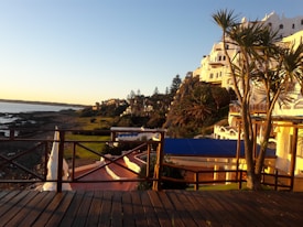 A coastal landscape featuring a white building complex perched on a hillside overlooking the ocean. The scene includes palm trees, a rocky shoreline, a wooden deck, and a pastel-colored sky at sunset. The architectural style is distinctive with curving shapes and bright colors, contrasting with the natural greenery and ocean.