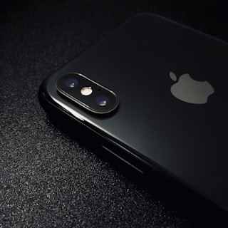 A close-up view of the back of a black smartphone with a dual camera setup and an Apple logo. The surface is smooth and reflective, highlighting the device's sleek design on a dark textured background.