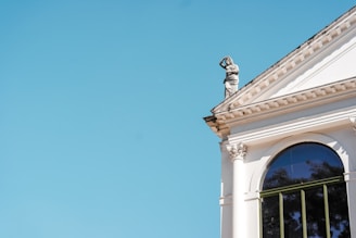 A classical building features ornate architectural details, including columns, a large arched window, and a statue on the roof. The sky is a clear, vibrant blue.