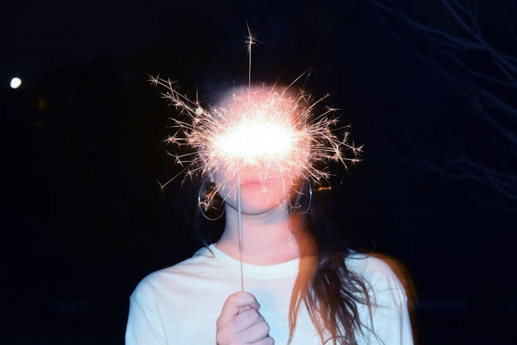 A photo of a woman holding a sparkler in front of her face.