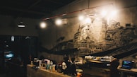 A dimly lit cafe or bar with a detailed mural on the wall depicting various architectural structures. There are several lights hanging overhead, an air conditioning unit, and coffee-making equipment on the counter. The atmosphere suggests a cozy, intimate setting.