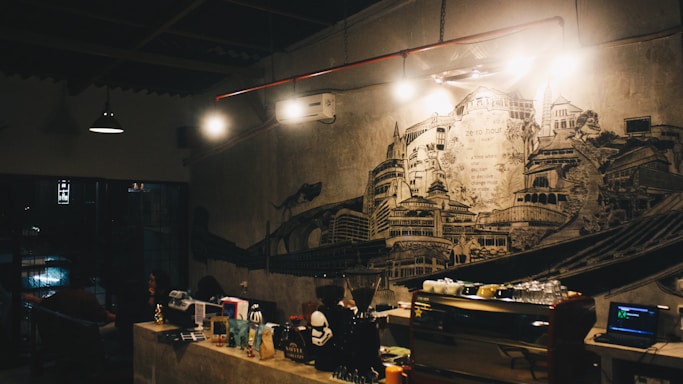A dimly lit cafe or bar with a detailed mural on the wall depicting various architectural structures. There are several lights hanging overhead, an air conditioning unit, and coffee-making equipment on the counter. The atmosphere suggests a cozy, intimate setting.