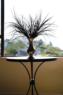 A brass vase with ornamental twigs or dried branches sits on top of a small round table. The table has slender, curved legs and stands against a bright, blurred outdoor background seen through a window.