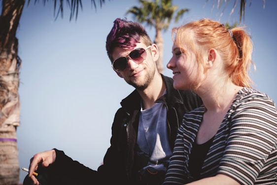 A man and a woman sitting outdoors under clear skies, surrounded by palm trees. The man wears sunglasses and has styled hair with a purple tint. The woman has red hair pulled back and is wearing a striped shirt. Both appear relaxed and engaged in a casual conversation.