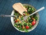 A fresh salad consisting of leafy greens, cherry tomatoes, black-eyed peas, and chopped bell peppers presented in a patterned bowl. A slice of whole grain bread rests on the side. A fork is placed strategically on top of the salad, all set against a textured dark blue table surface.