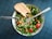 A fresh salad consisting of leafy greens, cherry tomatoes, black-eyed peas, and chopped bell peppers presented in a patterned bowl. A slice of whole grain bread rests on the side. A fork is placed strategically on top of the salad, all set against a textured dark blue table surface.