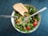 A fresh salad consisting of leafy greens, cherry tomatoes, black-eyed peas, and chopped bell peppers presented in a patterned bowl. A slice of whole grain bread rests on the side. A fork is placed strategically on top of the salad, all set against a textured dark blue table surface.