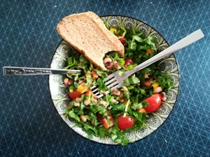 A fresh salad consisting of leafy greens, cherry tomatoes, black-eyed peas, and chopped bell peppers presented in a patterned bowl. A slice of whole grain bread rests on the side. A fork is placed strategically on top of the salad, all set against a textured dark blue table surface.