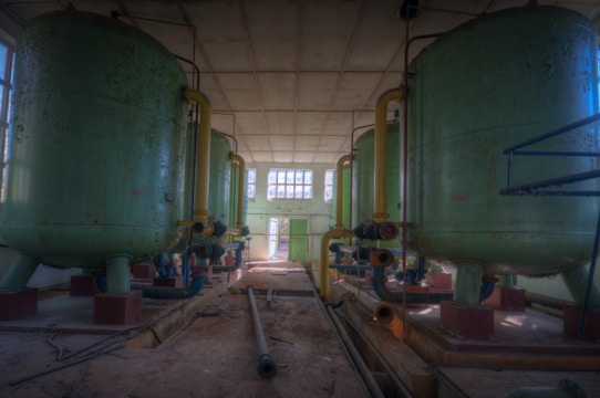 A spacious industrial room with large, cylindrical green tanks connected by yellow pipes. The tanks are elevated on sturdy legs and are situated on a dusty floor. Natural light enters through multiple windows at the far end, illuminating the interior. Several metal pipes and wires lie scattered haphazardly on the ground.