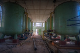 A spacious industrial room with large, cylindrical green tanks connected by yellow pipes. The tanks are elevated on sturdy legs and are situated on a dusty floor. Natural light enters through multiple windows at the far end, illuminating the interior. Several metal pipes and wires lie scattered haphazardly on the ground.