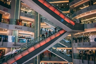 A multi-level shopping mall features several escalators crisscrossing through the center, with the surrounding floors filled with bright signage and shop displays. The environment appears busy with people moving up and down the escalators, and various store signs like 'Delicious', 'Playful', and 'Fashion' are visible.