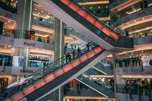 A multi-level shopping mall features several escalators crisscrossing through the center, with the surrounding floors filled with bright signage and shop displays. The environment appears busy with people moving up and down the escalators, and various store signs like 'Delicious', 'Playful', and 'Fashion' are visible.