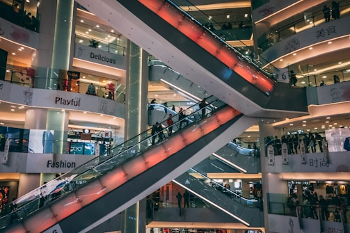 A multi-level shopping mall features several escalators crisscrossing through the center, with the surrounding floors filled with bright signage and shop displays. The environment appears busy with people moving up and down the escalators, and various store signs like 'Delicious', 'Playful', and 'Fashion' are visible.