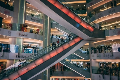 A multi-level shopping mall features several escalators crisscrossing through the center, with the surrounding floors filled with bright signage and shop displays. The environment appears busy with people moving up and down the escalators, and various store signs like 'Delicious', 'Playful', and 'Fashion' are visible.