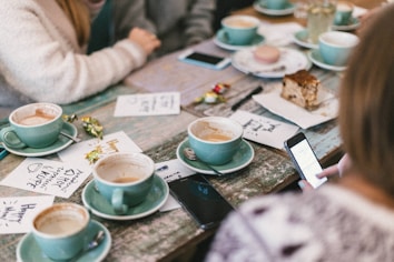 A wooden table is set with several light blue cups and saucers, some filled with coffee. There are handwritten notes on paper, colorful candy wrappers, a slice of cake, and smartphones. People are partially visible, sitting around the table, engaged in conversation.