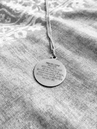 A round pendant with an engraved traveler's prayer sits on a textured fabric. The chain is visible, suggesting it's a necklace. The fabric has a subtle pattern, contrasting with the metallic surface of the pendant.