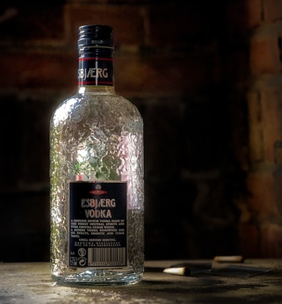 A clear, textured vodka bottle labeled 'Esbjerg Vodka' stands on a surface. The background is dimly lit, giving emphasis to the bottle's reflective surface. The atmosphere appears rustic, with a brick wall partially visible in the shadows.