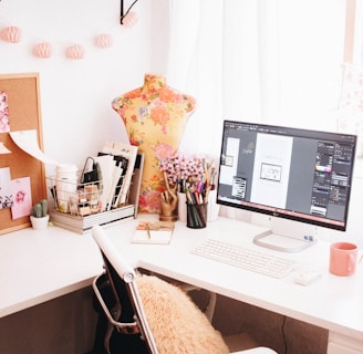 A bright, well-organized workspace featuring a computer displaying graphic design software. A floral-patterned dress form stands beside the monitor. A collection of pens and pencils is housed in a cup, along with a potted cactus and a wire basket containing books and a notebook. The desk chair is adorned with a fluffy cushion, and decorative string lights hang above.