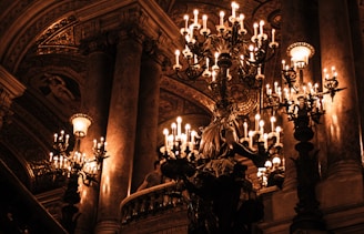 An opulent interior with ornate chandeliers casting a warm glow. The architecture features grand columns and intricate ceilings, creating a sense of luxury and elegance. The space feels historic with rich decorative elements.