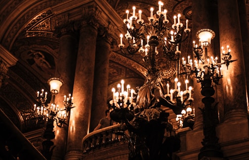 An opulent interior with ornate chandeliers casting a warm glow. The architecture features grand columns and intricate ceilings, creating a sense of luxury and elegance. The space feels historic with rich decorative elements.