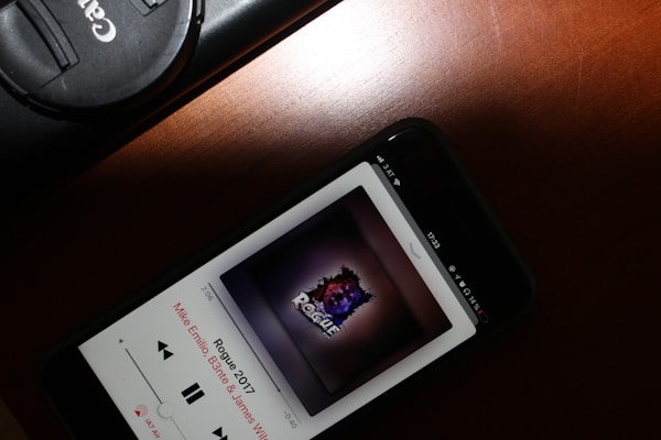 A smartphone placed on a wooden surface displays a music playing interface showing the album art for a song titled 'Rogue 2017' by Mike Emilio, B3nte & James Walshe. Next to the phone, there is a Canon camera lens cap.