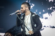 A man with short dark hair and sunglasses is performing on stage, holding a microphone. He is wearing a black jacket, a white shirt, and several necklaces. A person with long hair is visible in the background, blurred and in motion.