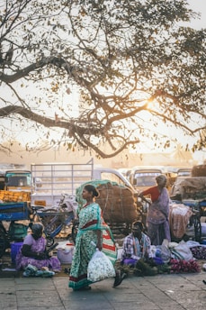 A street market scene featuring vendors sitting on the ground selling goods, with a large tree overhead. The early morning or late afternoon sunlight filters through the branches, casting a warm glow. Vehicles and stalls are in the background, and a woman in a green dress carries a bag, walking past.