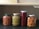 Four jars containing preserved fruits and vegetables are neatly arranged on a dark surface. The jars contain small round fruits, sliced green vegetable stalks, red berries, and sliced yellow fruits, each with different colored lids.