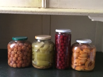 Four jars containing preserved fruits and vegetables are neatly arranged on a dark surface. The jars contain small round fruits, sliced green vegetable stalks, red berries, and sliced yellow fruits, each with different colored lids.