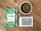 A notebook with a green leaf pattern cover featuring the text 'AHORA es el momento' is placed on a wooden surface alongside a small round cactus in a white pot and an e-reader displaying text.