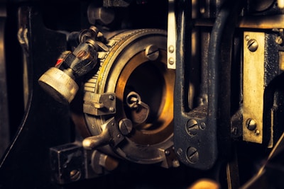 A close-up of an intricate mechanical device with various metal components, including a dial with numbers, screws, and levers. The metallic parts exhibit a mix of silver and gold tones, with some visible wear and tear, giving it an industrial and vintage appearance.