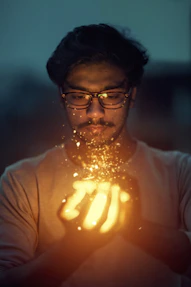 A man with glasses gazes intently at a bright, glowing light held between his hands. The light emits sparkling particles that illuminate his face against a dim, blurred background.