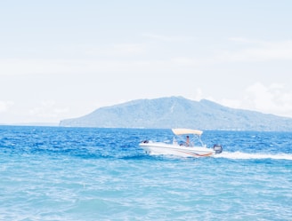 A speedboat glides across a bright blue ocean, with white spray following behind it. The background features a distant, lush mountain under a clear sky.