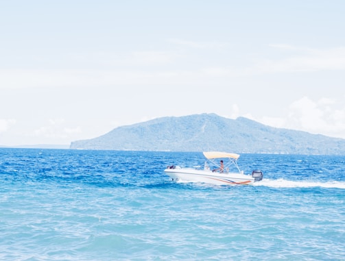 A speedboat glides across a bright blue ocean, with white spray following behind it. The background features a distant, lush mountain under a clear sky.