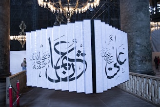 A large installation of vertical panels displaying intricate Arabic calligraphy stands in an ornate, historical interior space. The panels are arranged in a fan-like structure with a chandelier above, casting light throughout the setting. The area is bordered by grand marble columns and decorative railings.