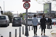 A busy street scene with people walking along a sidewalk, passing by a speed limit sign indicating a maximum speed of 30. Cars are parked and moving on the road. In the background, there are several commercial signs and a crowd near what appears to be a ferry terminal.