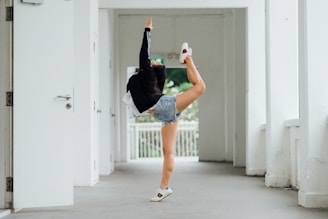 A person performing a high kick in a hallway, with one leg extended and the other grounding their stance. The person wears a black jacket, denim shorts, and white sneakers. The background features white walls and doors, with a railing and greenery visible at the far end.